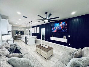 Ein modernes Wohnzimmer mit einem großen Fernseher, einem Deckenventilator und einer Küche im Hintergrund.