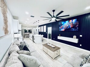 Ein modernes Wohnzimmer mit einem großen Fernseher, einem Deckenventilator und einer Küche im Hintergrund.