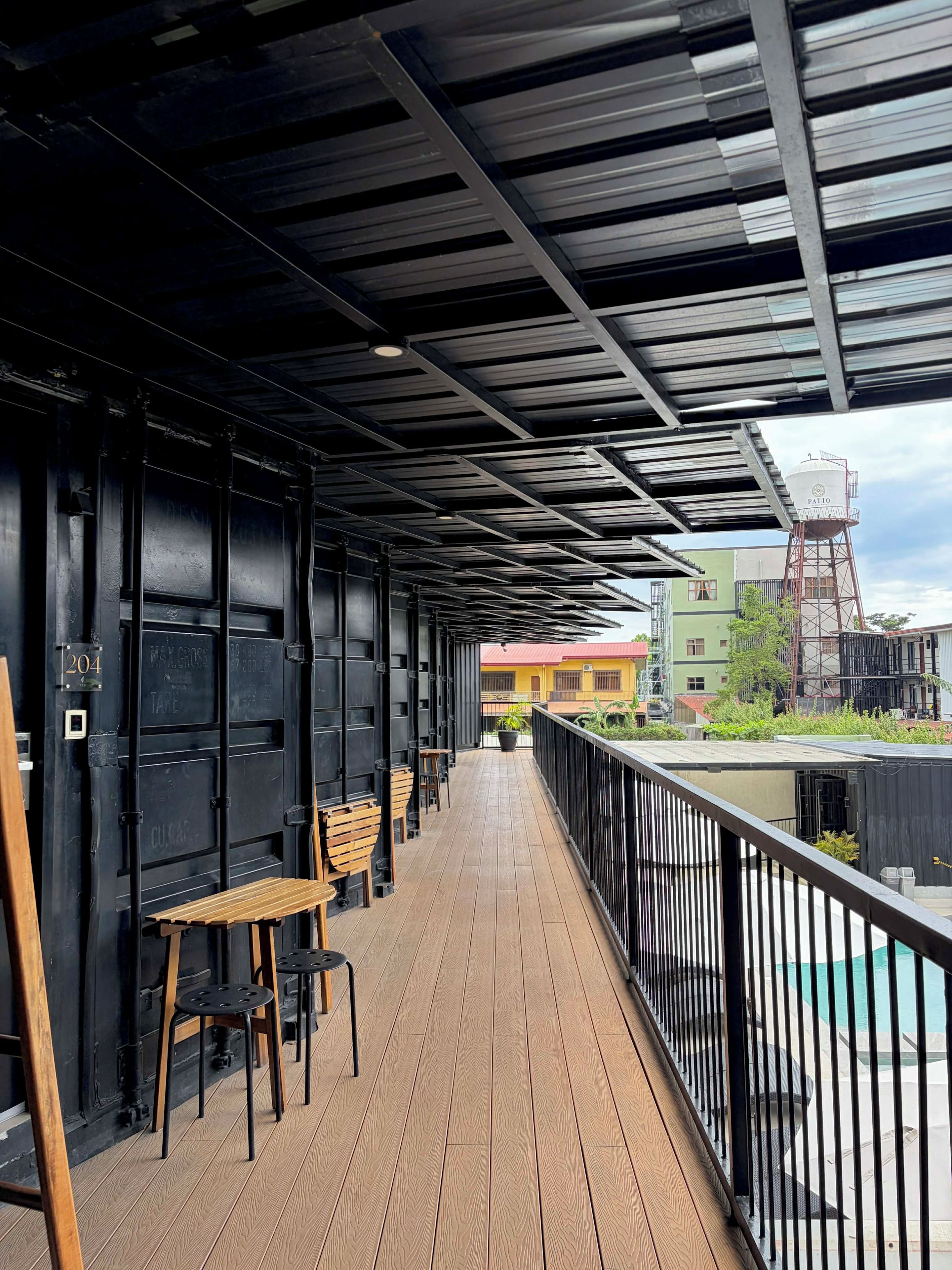 A covered outdoor area with a wooden deck, black metal railings, and a view of buildings in the background.