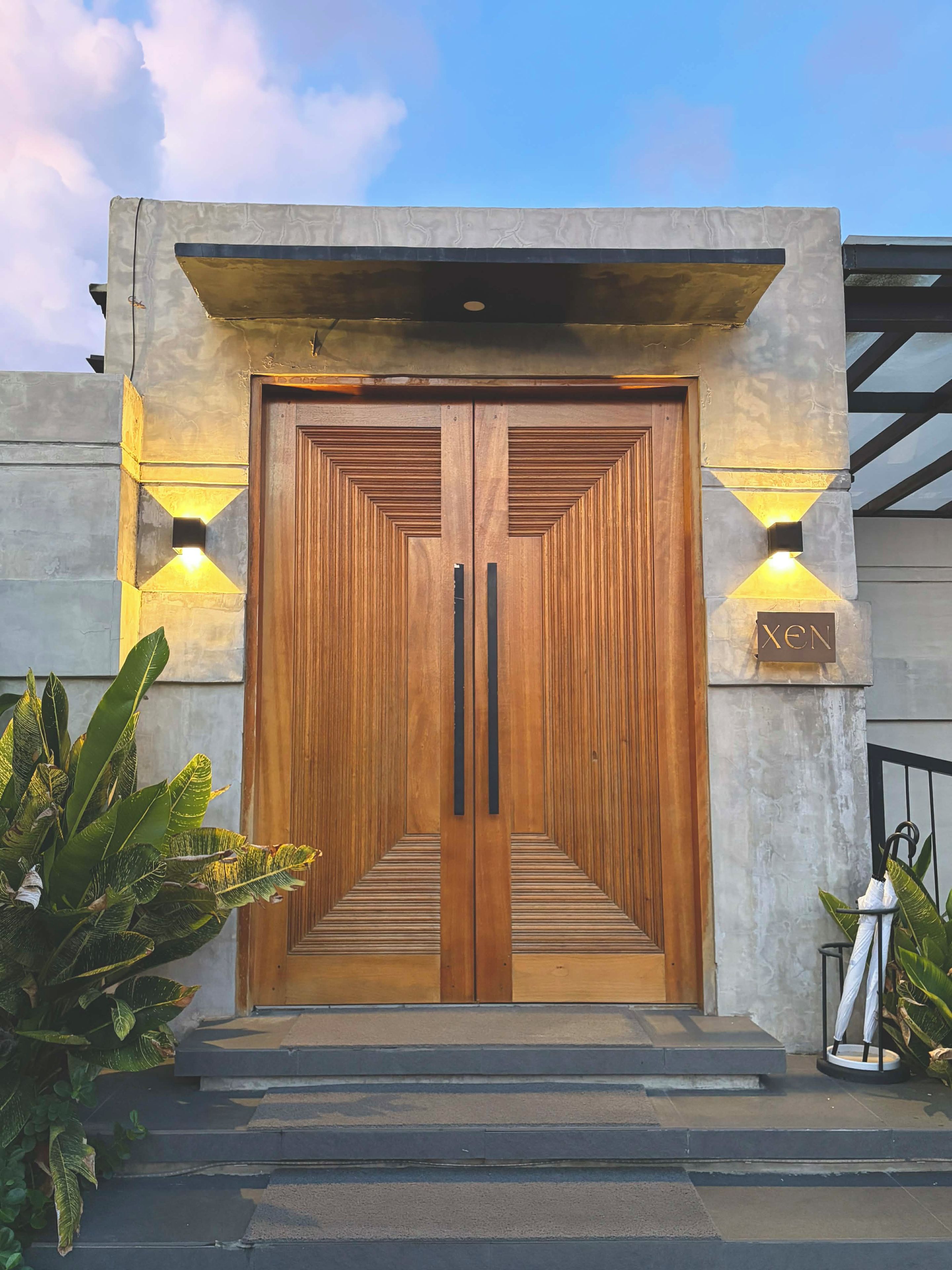 A double wooden door with a geometric pattern, flanked by two wall-mounted lights, and a sign above the door.