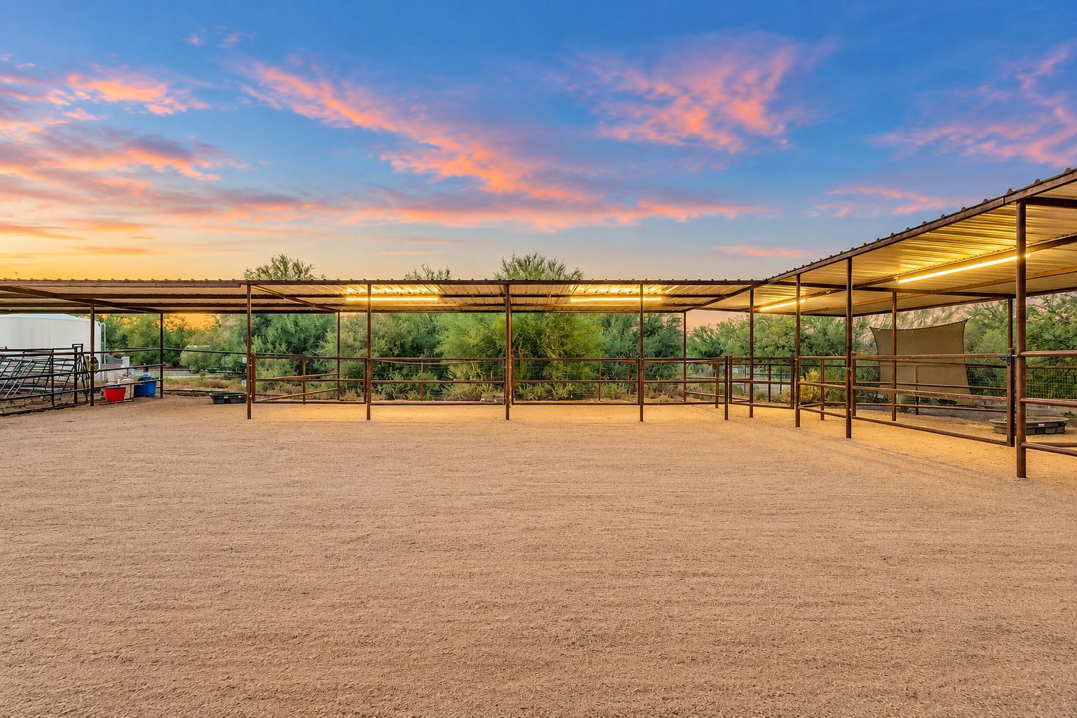 An outdoor equestrian arena with a metal roof structure, surrounded by greenery.