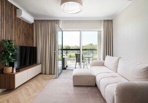 A modern living room with a flat-screen TV, a beige sofa, a wooden floor, and a large window with curtains. - Lotnisko Chopina Luxury Apartment (Warszawa)