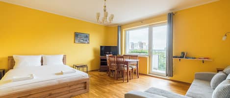 Habitación de hotel con cama, televisión, mesa de comedor con sillas, sofá y ventana grande con vista a edificios.