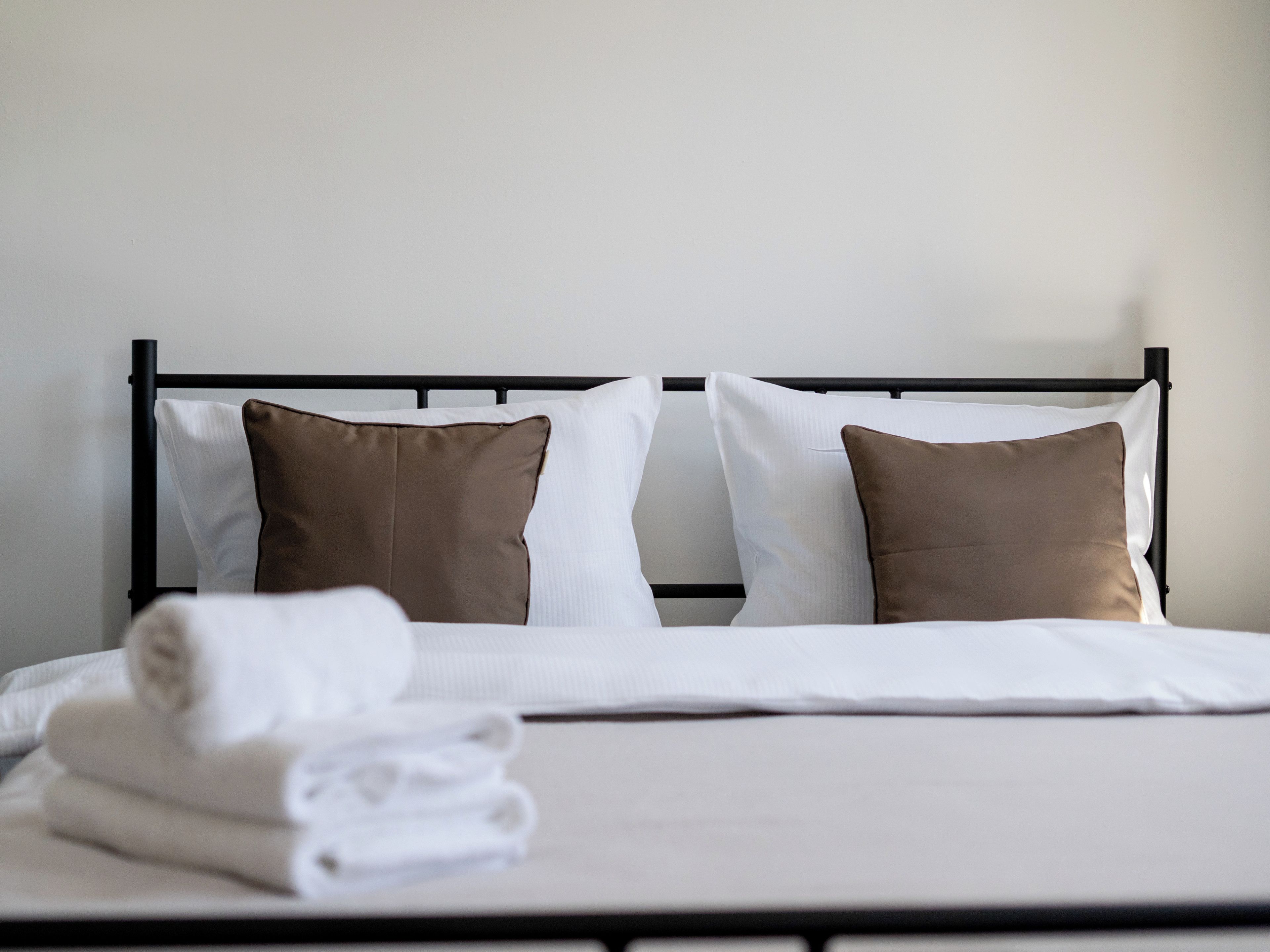 Cama com estrutura metálica preta, roupa de cama branca e duas almofadas castanhas.