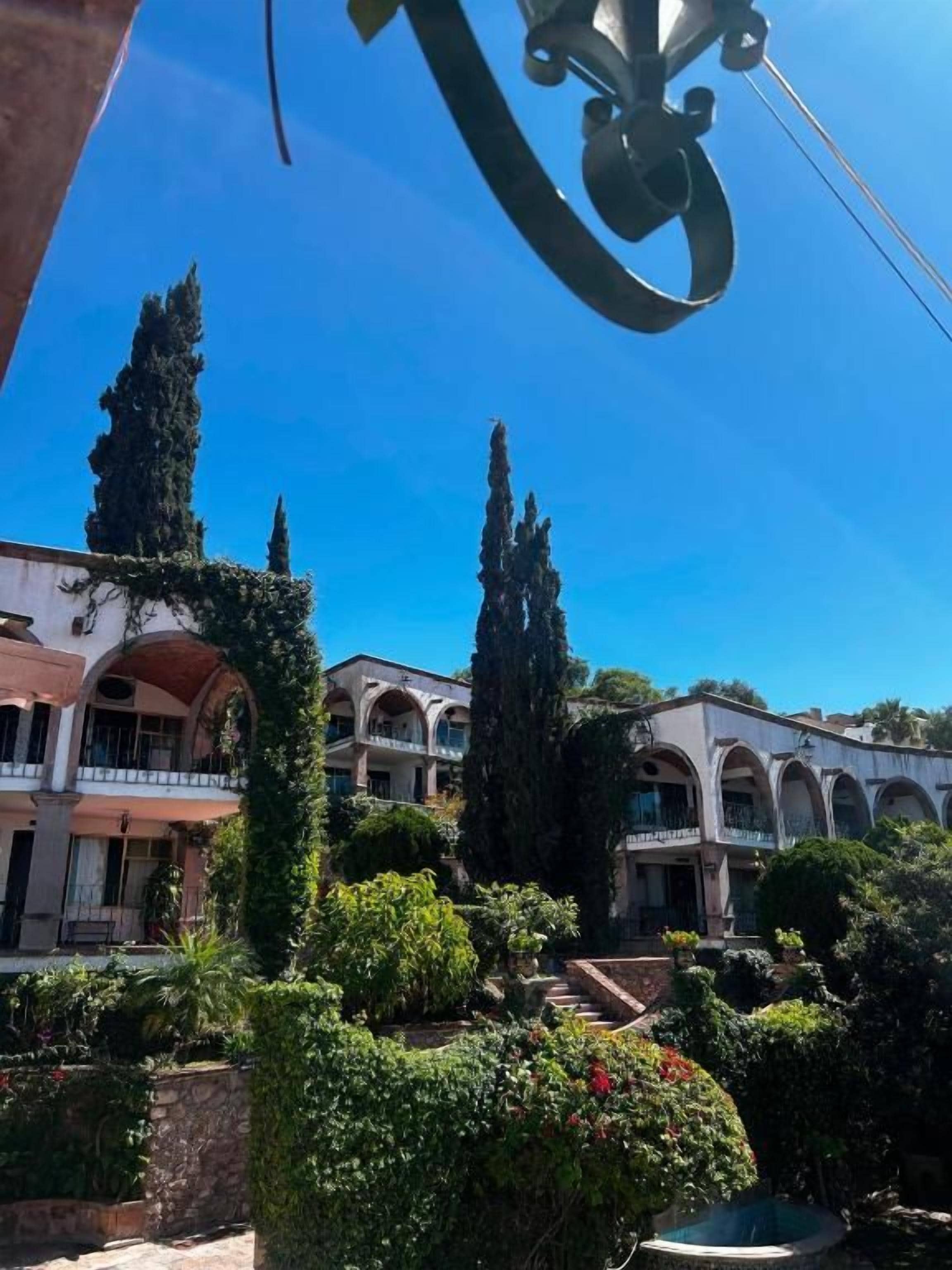 Un patio con una fuente, rodeado de follaje y altos cipreses, con un edificio con arcos al fondo.