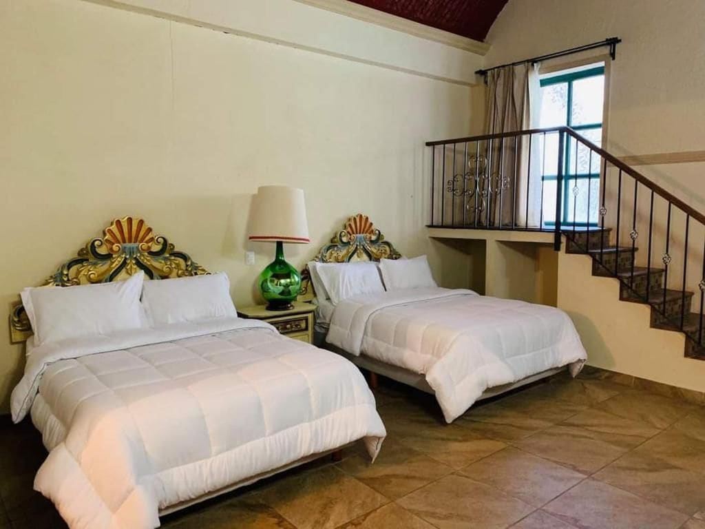 Un dormitorio con dos camas, una mesita de noche con lámpara, una escalera y un ventanal con cortinas.