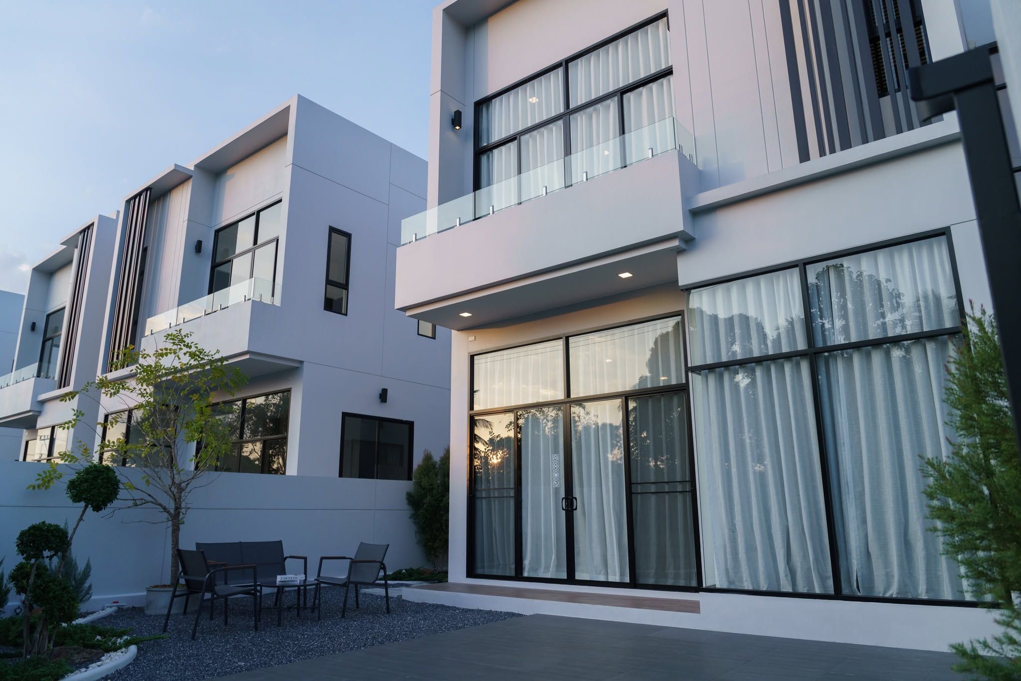 A modern residential building with a flat facade, large windows, and a small outdoor seating area with chairs.