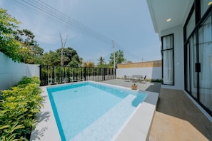 Un área de piscina moderna al aire libre con una piscina azul cristalina, rodeada por una valla baja, y contigua a un edificio con amplias ventanas.