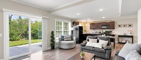 A spacious living area with a large sliding glass door leading to a patio, a kitchen with dark cabinets and a stone backsplash, and a seating area with a gray sofa and a coffee table.