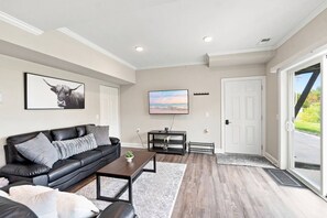 A modern living room with a black leather sofa, a coffee table, a flat-screen TV, and a door leading to a covered outdoor area. - Spacious Modern 4BR/4BA First-Floor Accessible (Eggertsville)