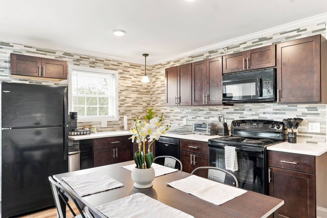A modern kitchen with dark wood cabinets, a black refrigerator, a microwave, and a stove. There is a window with blinds, a vase with flowers on the counter, and a dining table set with placemats.