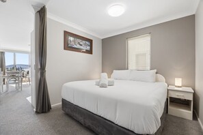 Ein Hotelzimmer mit einem großen Bett, einem Nachttisch mit Lampe, einem Fenster mit Jalousien, einem gerahmten Bild an der Wand und Blick ins Freie.
