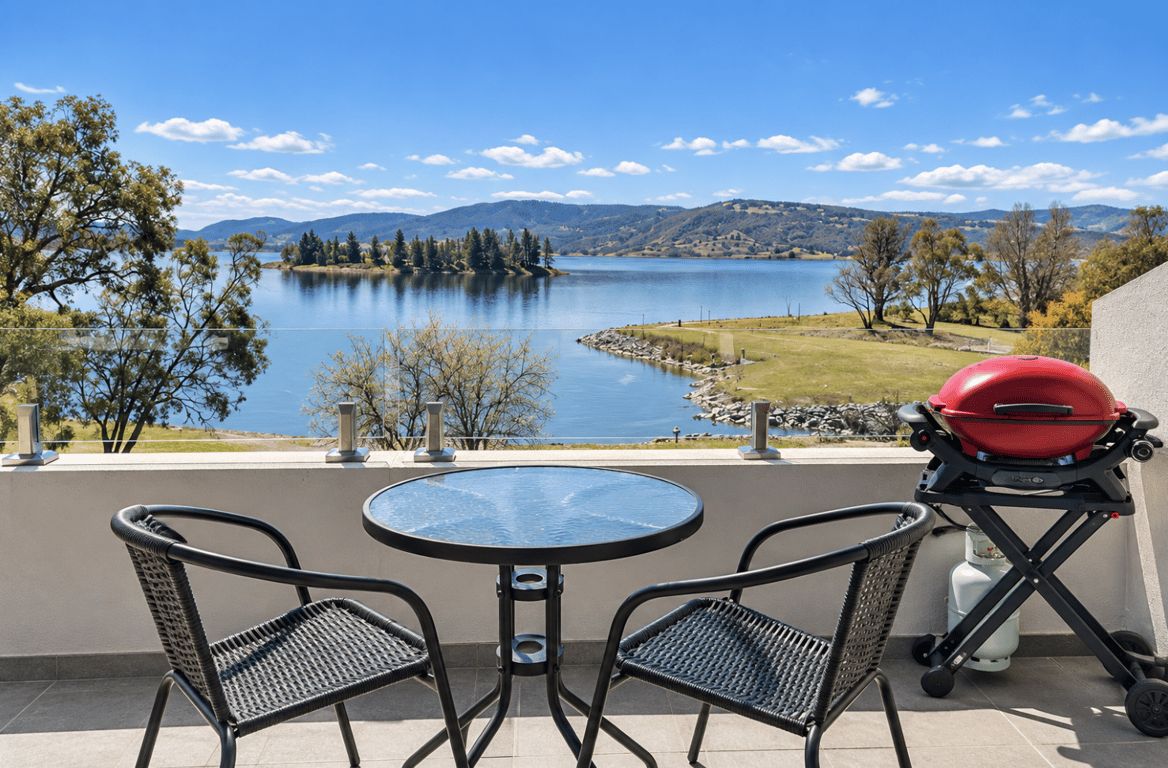 A balcony with a round table and two chairs, overlooking a lake with mountains in the background.