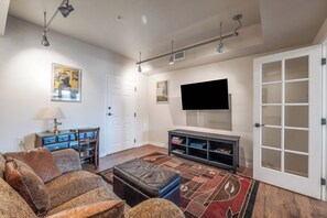 A living room with a sofa, ottoman, entertainment unit, and a flat-screen TV mounted on the wall. - Gated Riverfront Condo W/ Private Boardwalk (Post Falls)