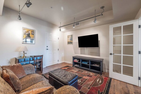 Un salon comprenant un canapé, un pouf, un meuble de rangement pour appareils électroniques et un téléviseur à écran plat fixé au mur.
