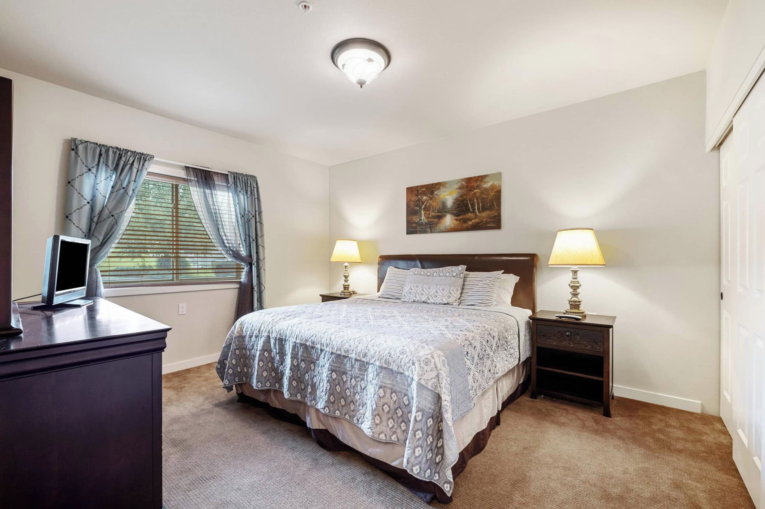 A bedroom with a bed, two bedside lamps, a painting on the wall, a desk with a computer, and a window with curtains.