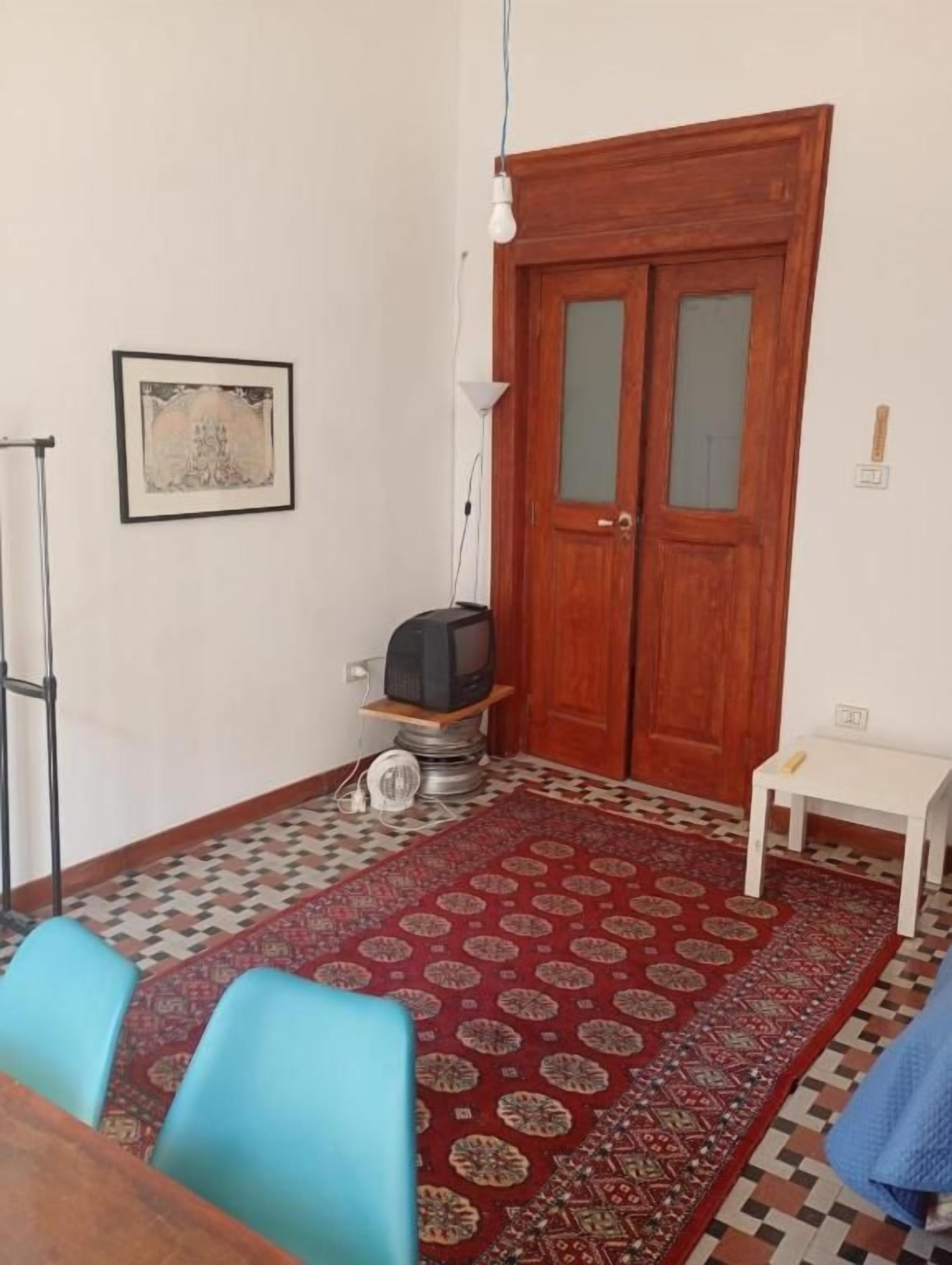 Une pièce avec une porte en bois, un téléviseur posé sur un meuble, un tapis à motifs et une petite table blanche.
