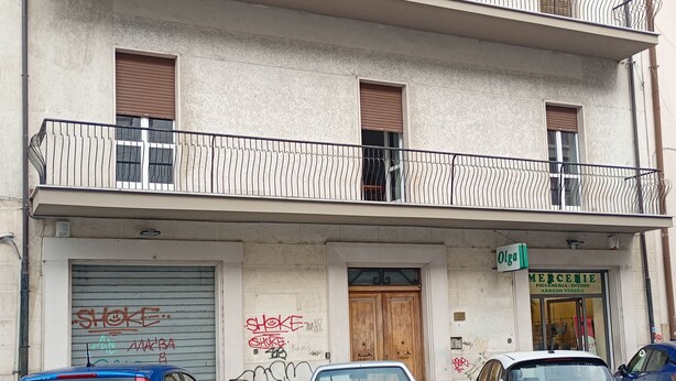 A building with a closed shutter, graffiti, and parked cars. - Roma 64 (Campobasso)
