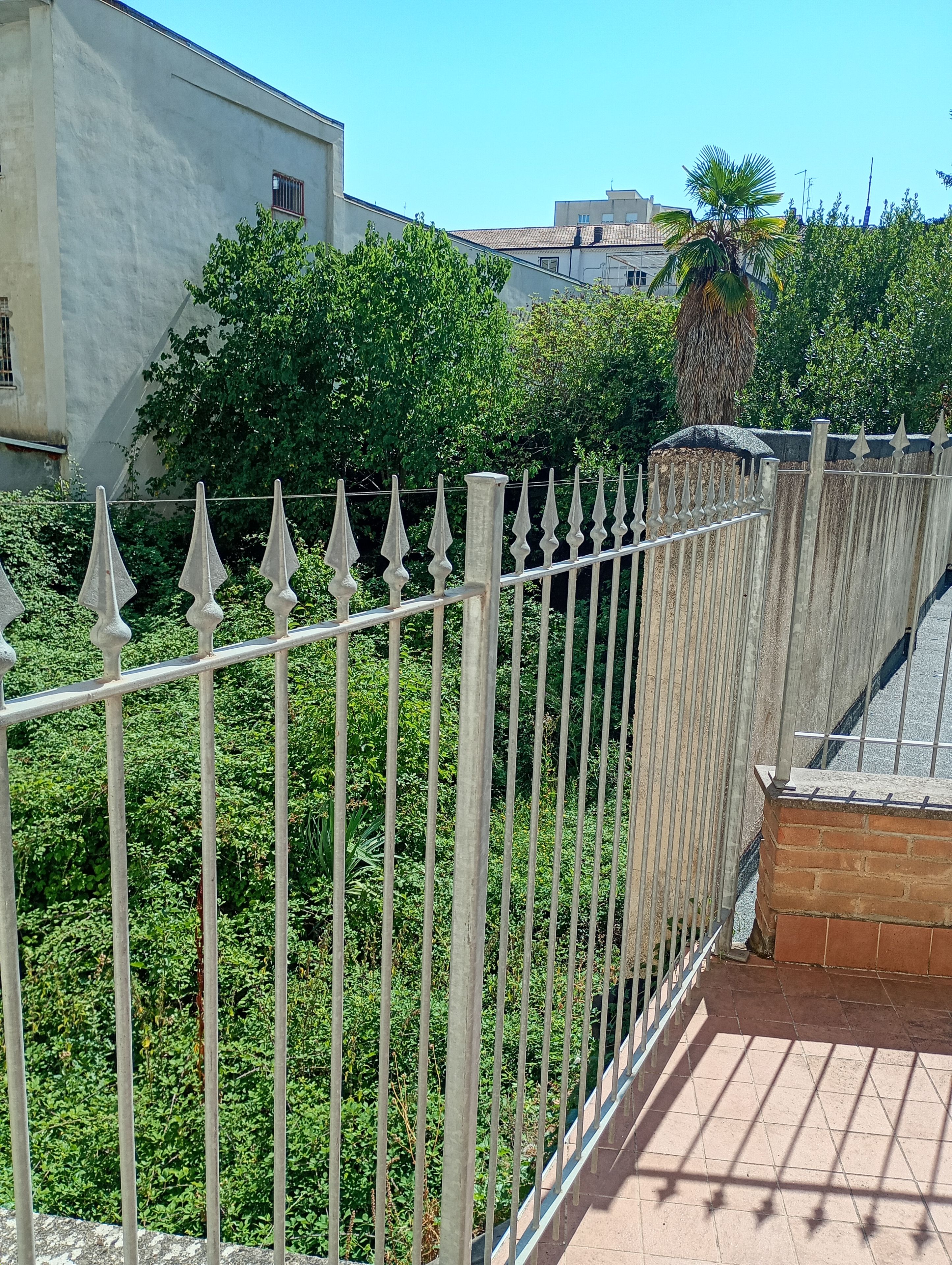 Una valla de metal con remates puntiagudos, un muro de ladrillo y abundante follaje verde al fondo.
