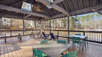 An outdoor covered patio with tables and chairs, a wooden deck, and a view of a forested area.
