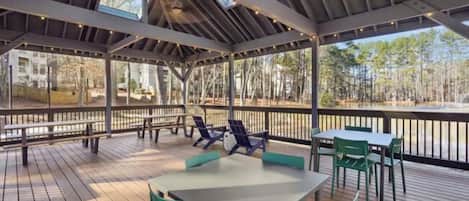 An outdoor covered patio with tables and chairs, a wooden deck, and a view of a forested area.