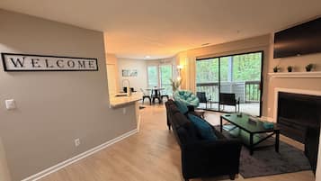 A modern living room with a black sofa, a coffee table, and a fireplace. There is a 'WELCOME' sign on the wall and a dining area in the background.
