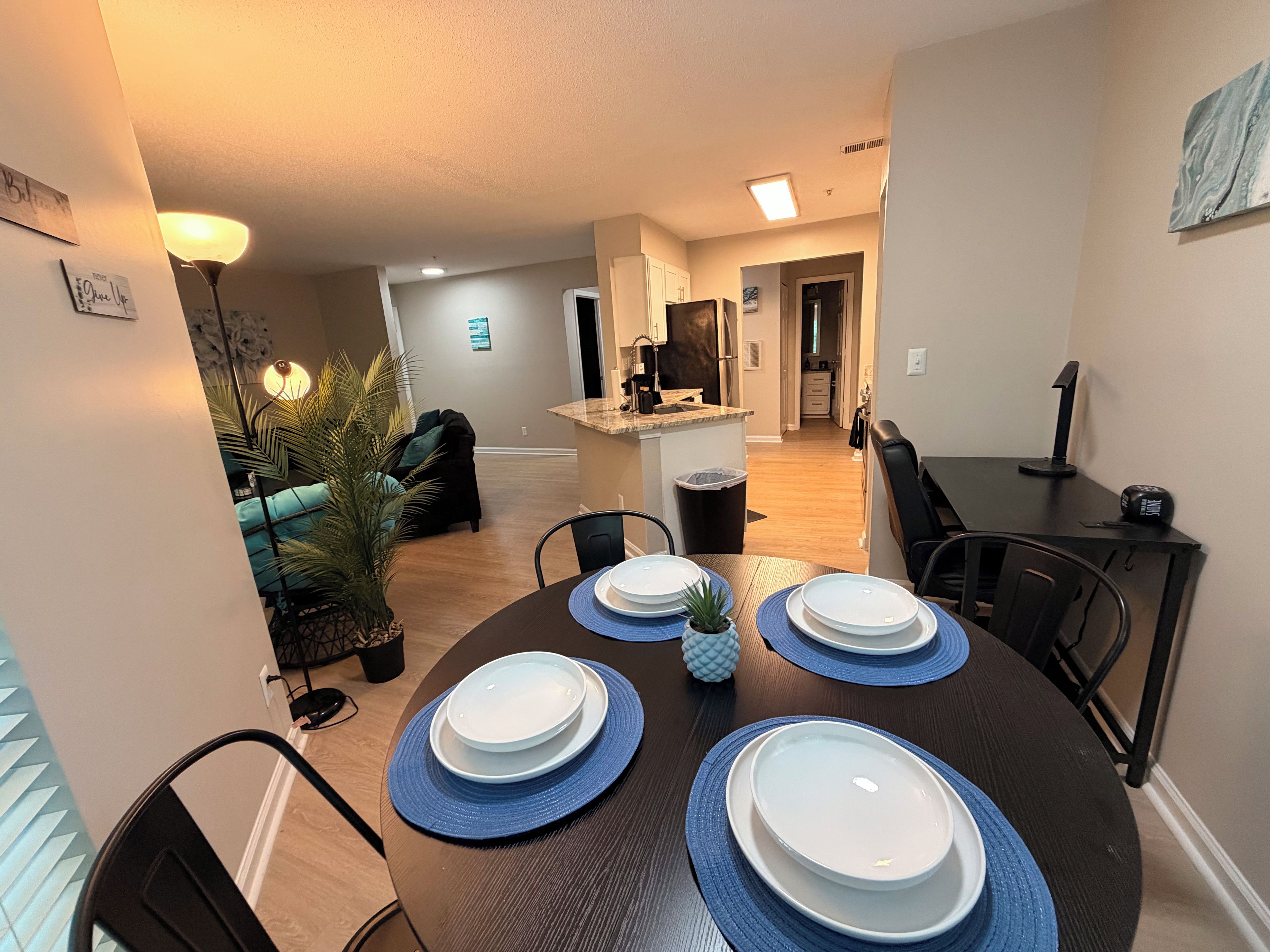 A dining area with a round table set for four, a kitchen in the background, and a living room with a couch and a plant.