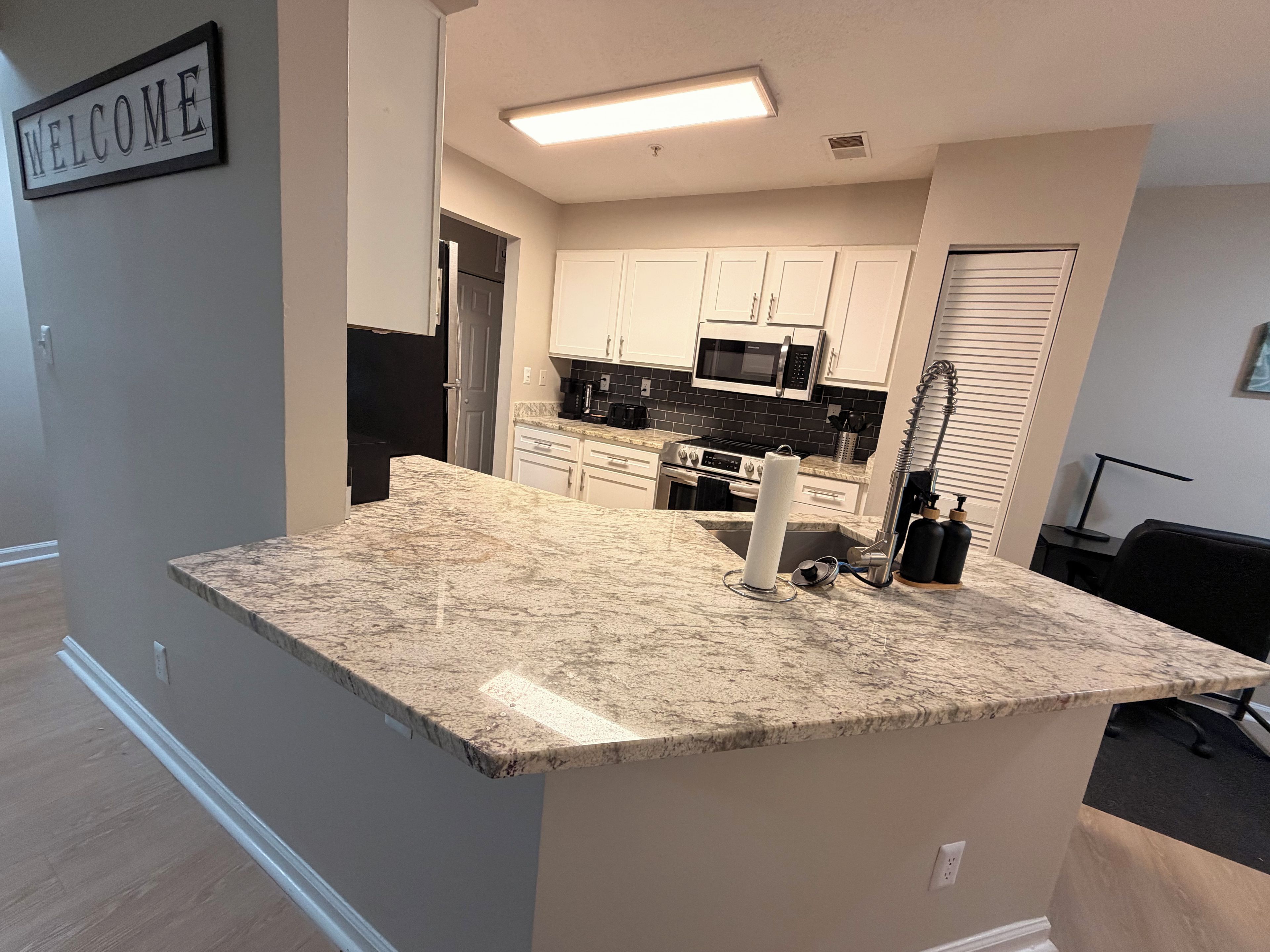 A modern kitchen with white cabinets, a granite countertop, and a welcome sign.