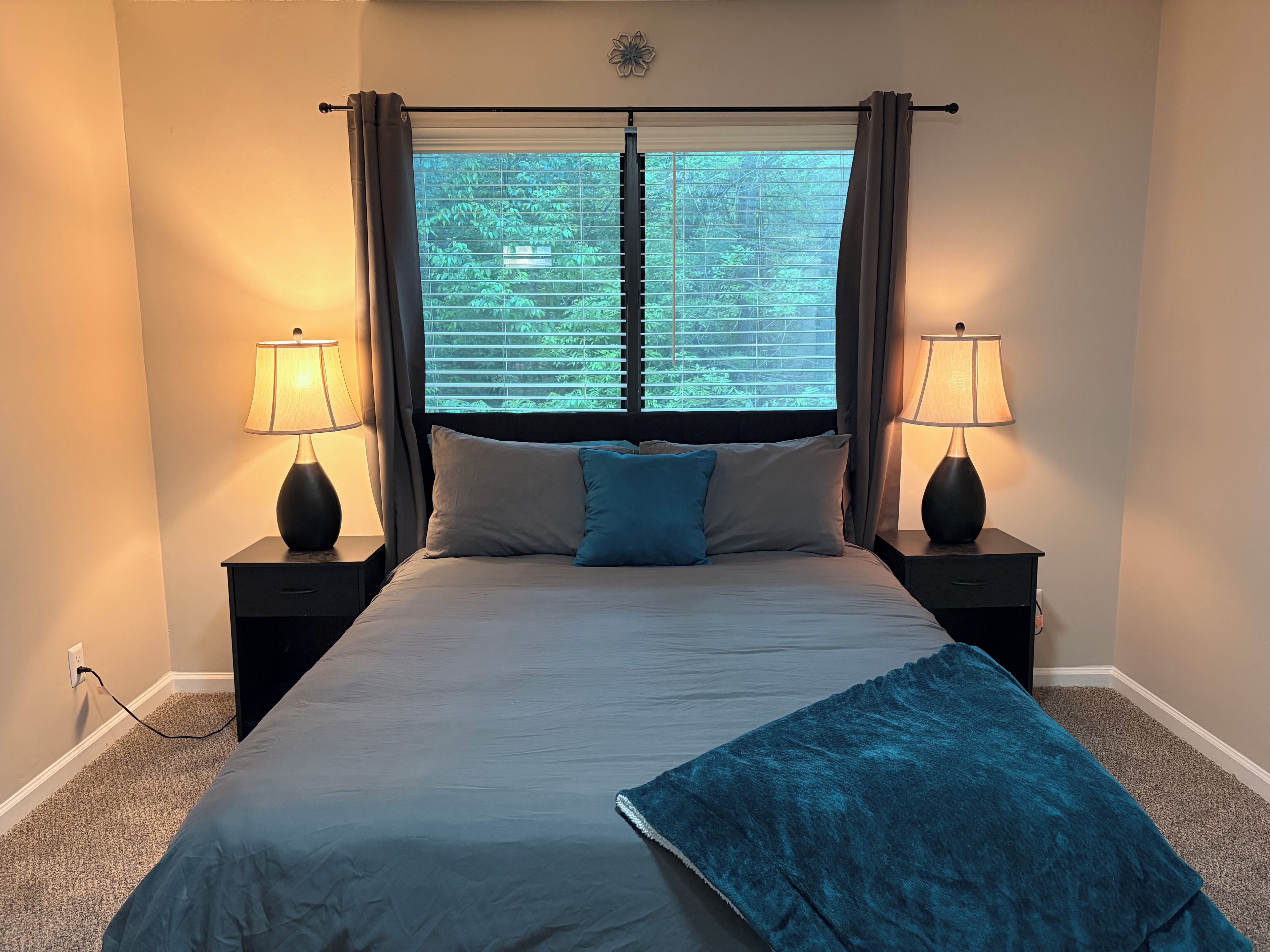 A bedroom with a bed, two bedside lamps, a window with blinds, and a view of greenery outside.