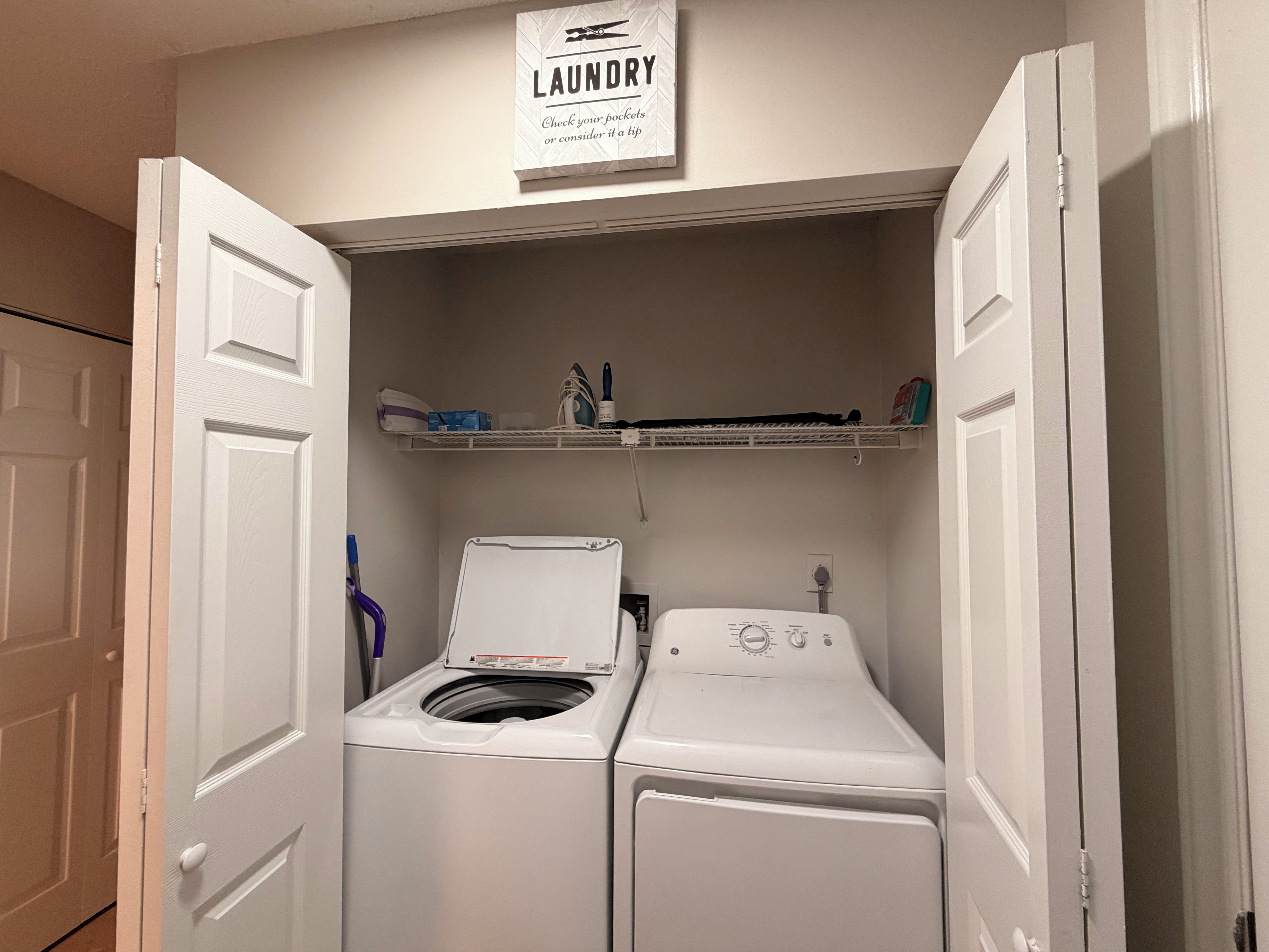 A laundry room with a washing machine and dryer, a shelf with laundry supplies, and a sign above the door.