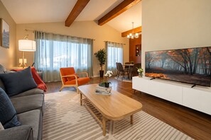 A modern living room with a sofa, armchair, coffee table, and a TV. There is a dining area with chairs and a table, and a large window with curtains. - Bright Family Home | 10 mins to Airport & Downtown (Calgary)