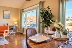 Un coin repas avec une table dressée pour deux personnes, une chaise, une plante et un vase rempli de fleurs.