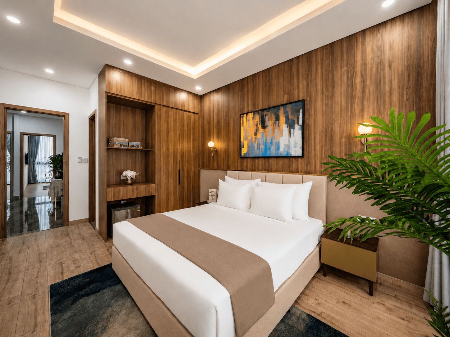 Una habitación de hotel moderna con una cama grande, paneles de madera, un cuadro, una mesita de noche y una planta en maceta.