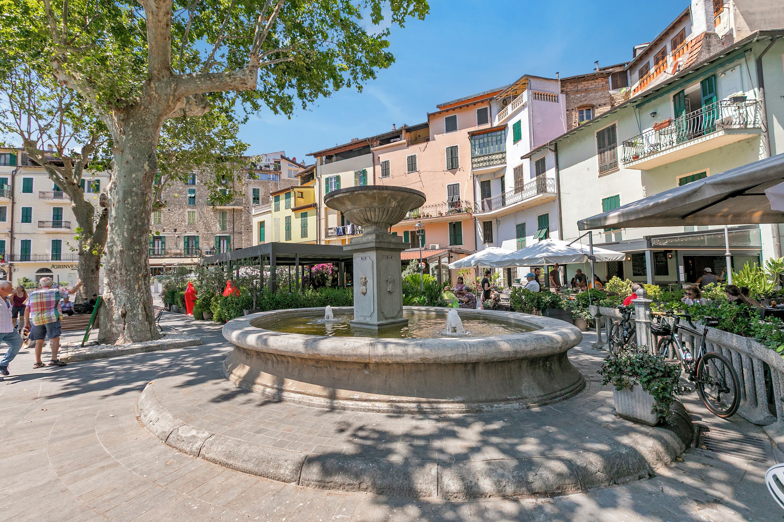 A public square with a central fountain, surrounded by multi-story buildings with balconies and outdoor seating areas.
