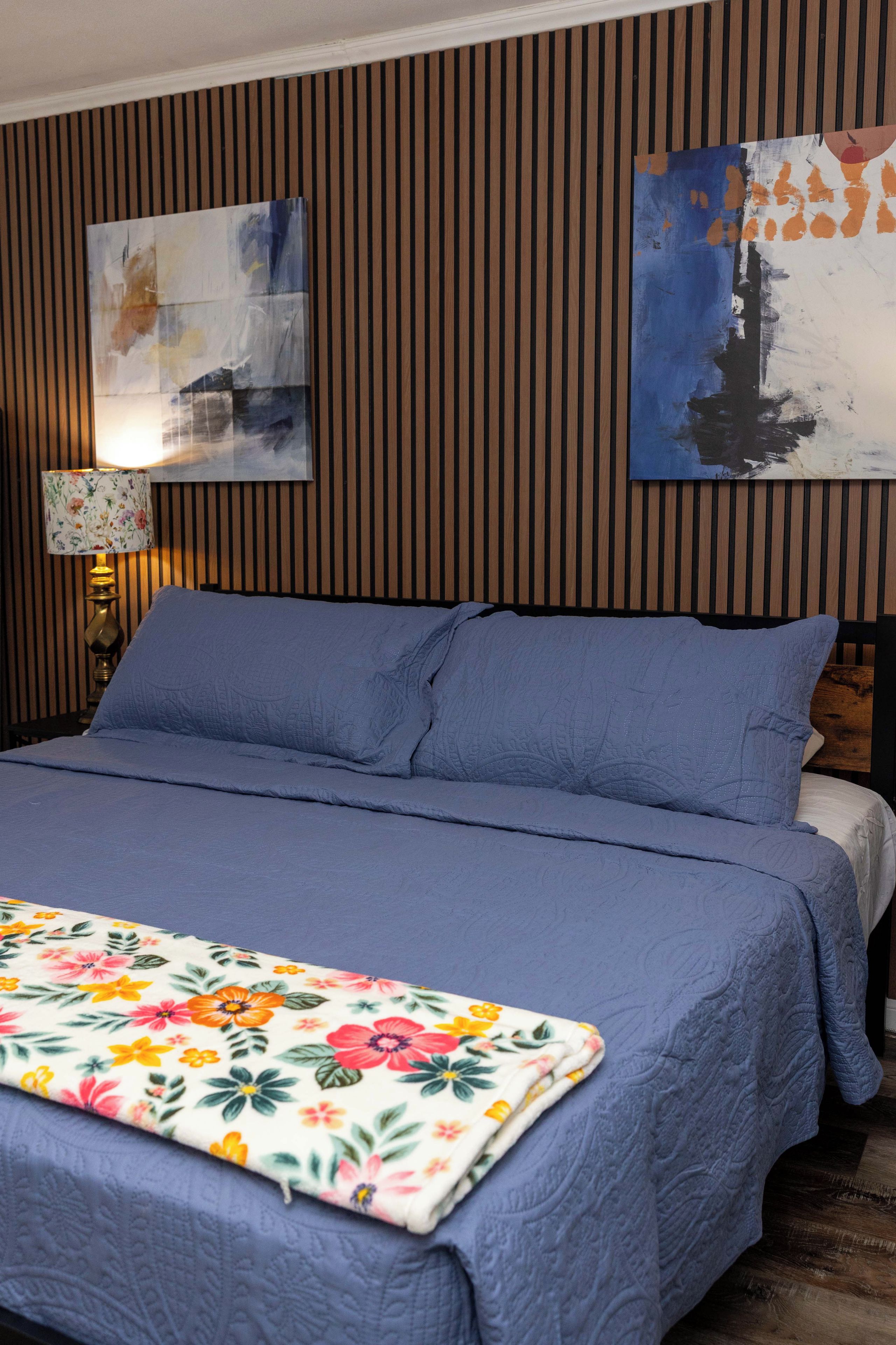 Un lit avec une couette bleue et des oreillers, un plaid à motifs floraux, une lampe et deux tableaux accrochés au mur.