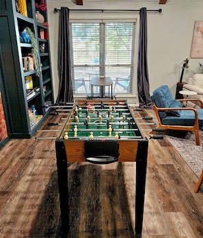 A room with a foosball table, a bookshelf, a window with curtains, and a sofa.