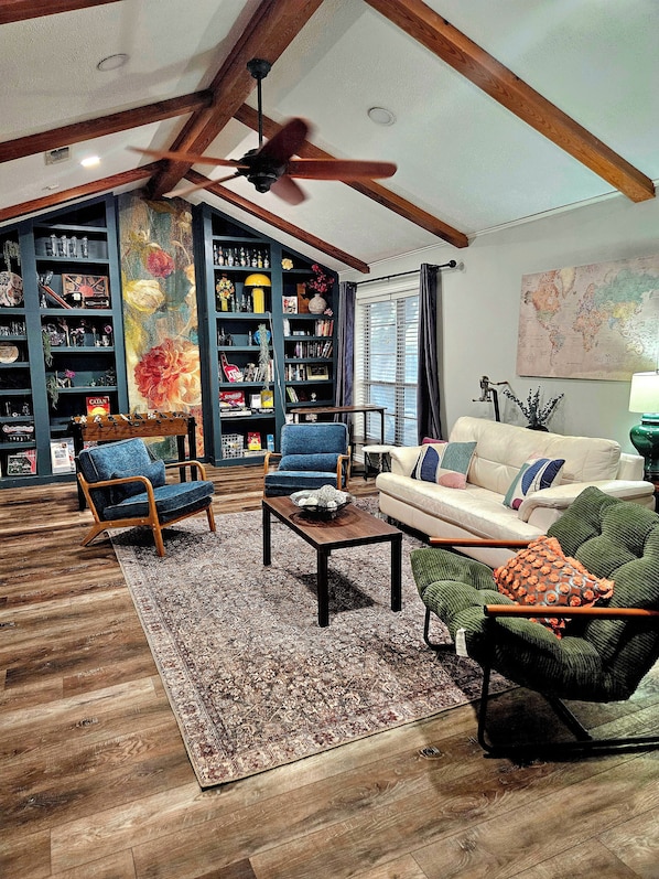A spacious living room with a high ceiling, wooden beams, a ceiling fan, a large bookshelf, a coffee table, and a sofa.