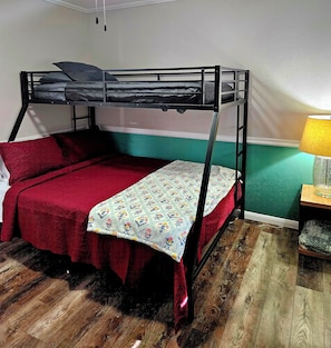 A bunk bed with a red bedspread and a patterned sheet, a wooden floor, and a lamp on a side table.