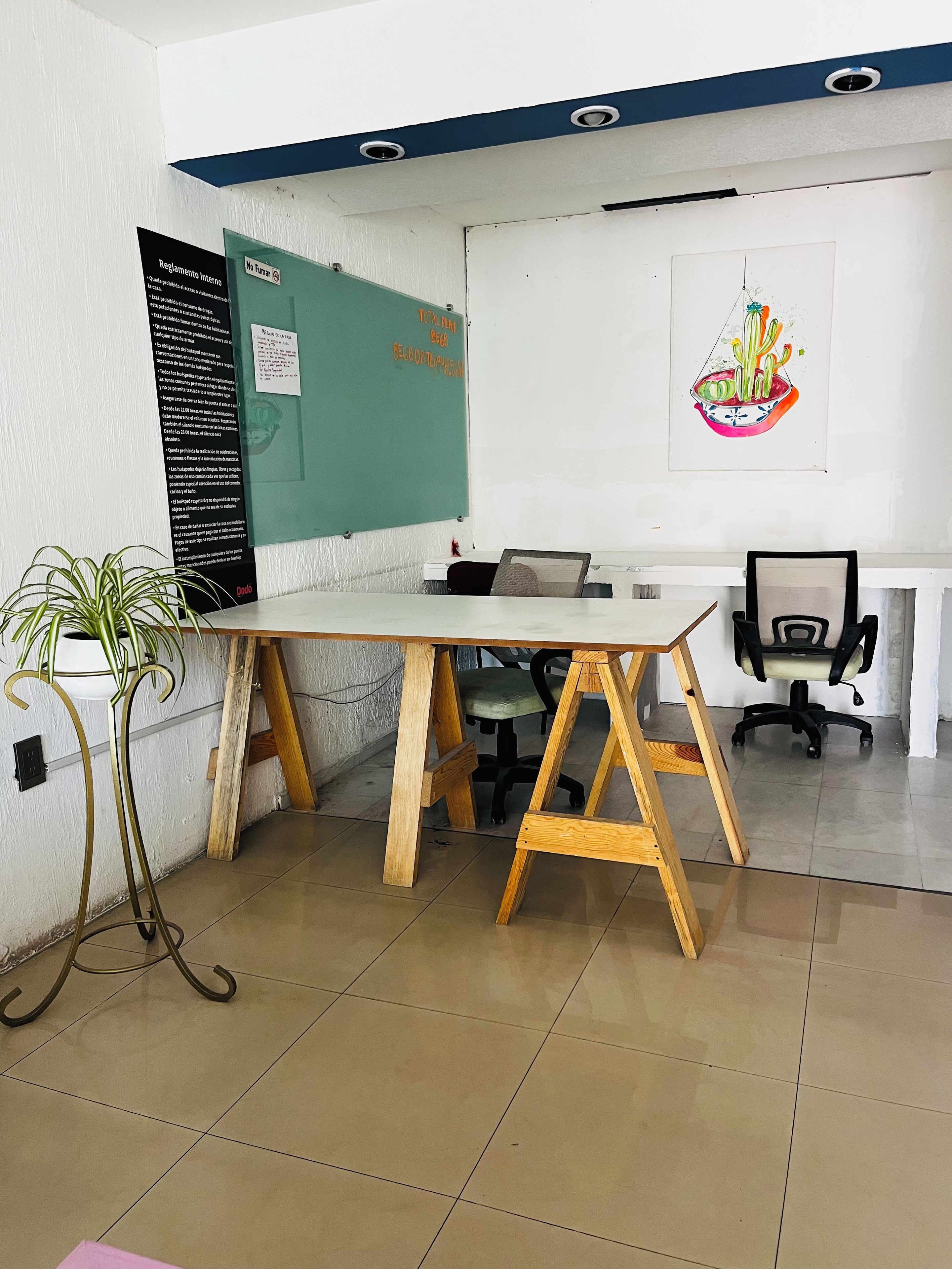 A conference room with a whiteboard, a green bulletin board, a desk with a chair, and a potted plant.