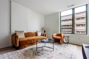 Ein Wohnzimmer mit einer Couch, einem kleinen runden Tisch, einem Sessel und einem großen Fenster mit Blick auf ein Gebäude.