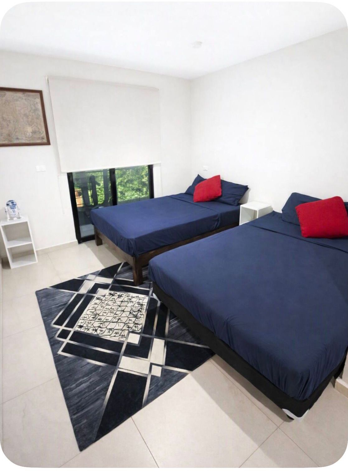 A modern bedroom with two beds, a geometric patterned rug, and a window with a view of greenery.