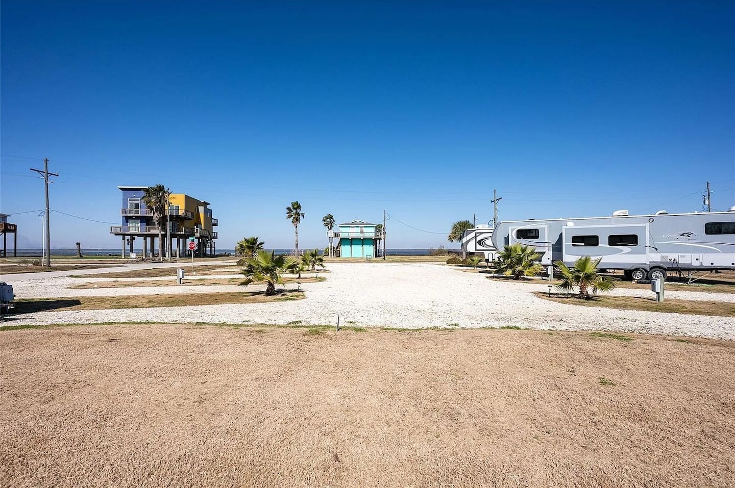 Un parque para vehículos recreativos frente a la playa, con cielo despejado, un edificio con balcón y palmeras.