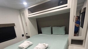 A compact room with a bunk bed, a small window with blinds, a bedside table, and a trash bin.