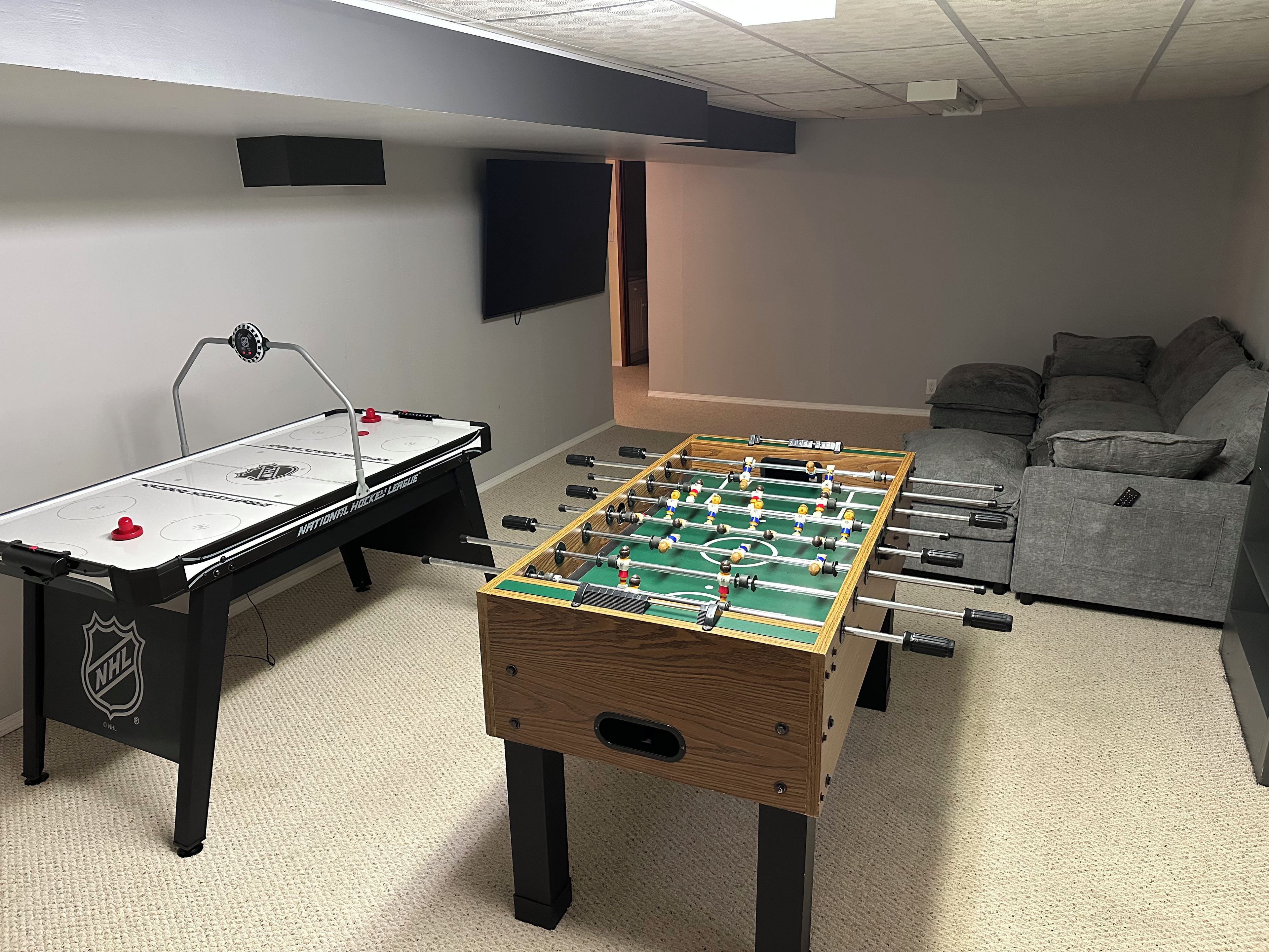 Um quarto com duas mesas de futebol de mesa, um sofá cinza e uma TV de tela plana presa à parede.