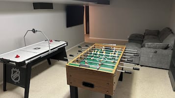 Um quarto com duas mesas de futebol de mesa, um sofá cinza e uma TV de tela plana presa à parede.