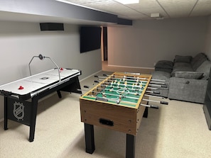 A room with two foosball tables, a grey sofa, and a flat-screen TV mounted on the wall.