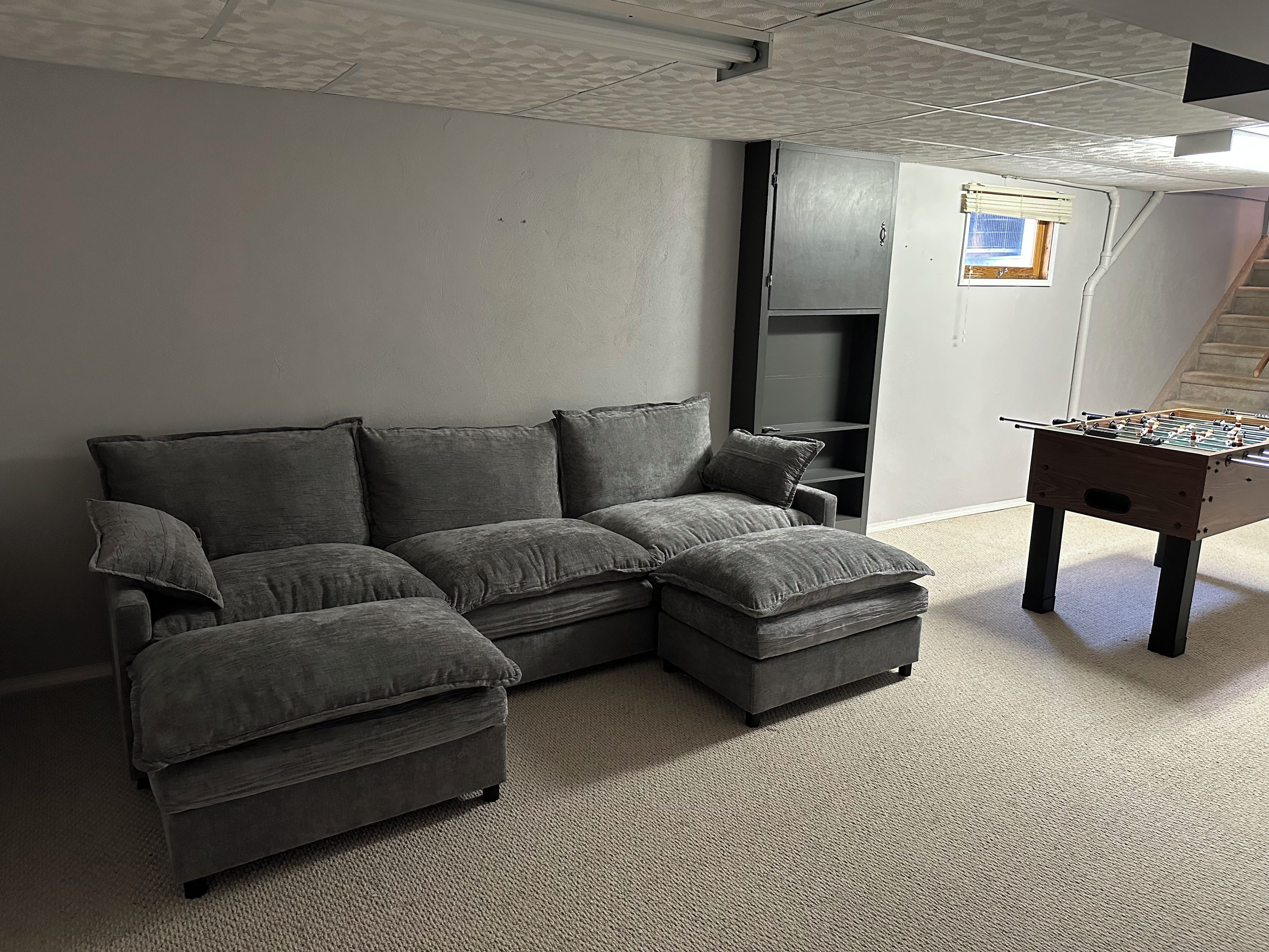 Um sofá modular cinza com um banquinho que combina, uma mesa de futebol e uma escada ao fundo.