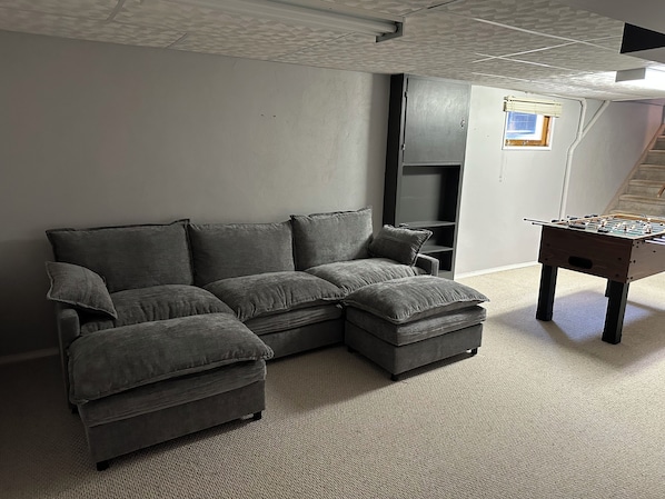 A grey sectional sofa with a matching ottoman, a foosball table, and a staircase in the background.