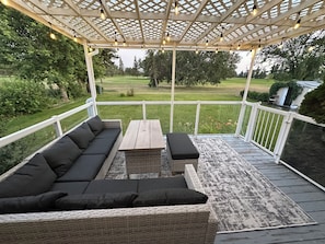 A covered outdoor seating area with a sofa, table, and bench, overlooking a grassy yard.