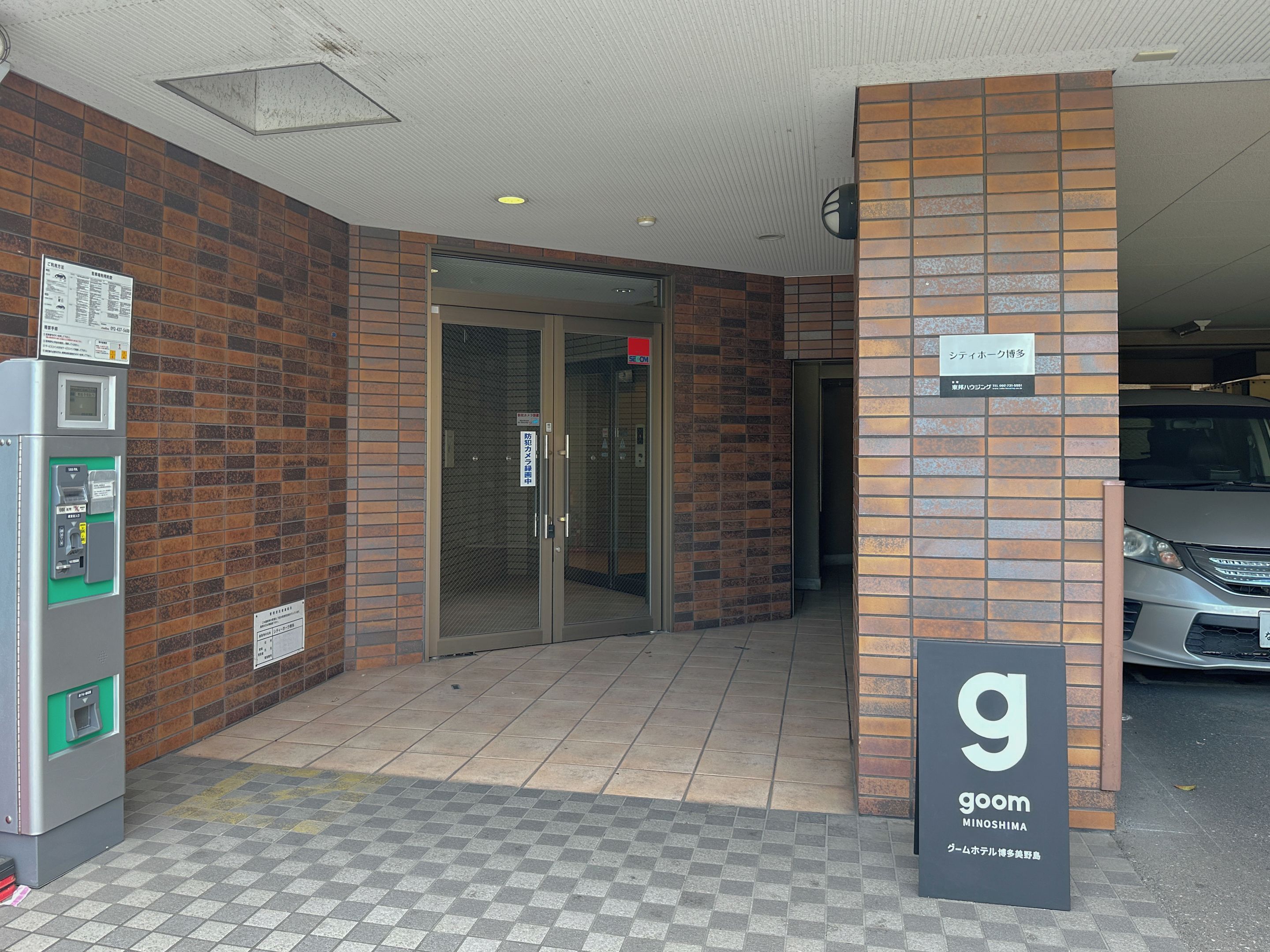 A building entrance with a vending machine, a glass door, and a sign for 'goom MINOSHIMA'.